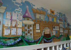 Graffiti zamku na ścianie w szatni przedszkolnej a na nim dyplomy w srebrnych ramkach, otrzymane przez przedszkole za udział dzieci w licznych konkursach.